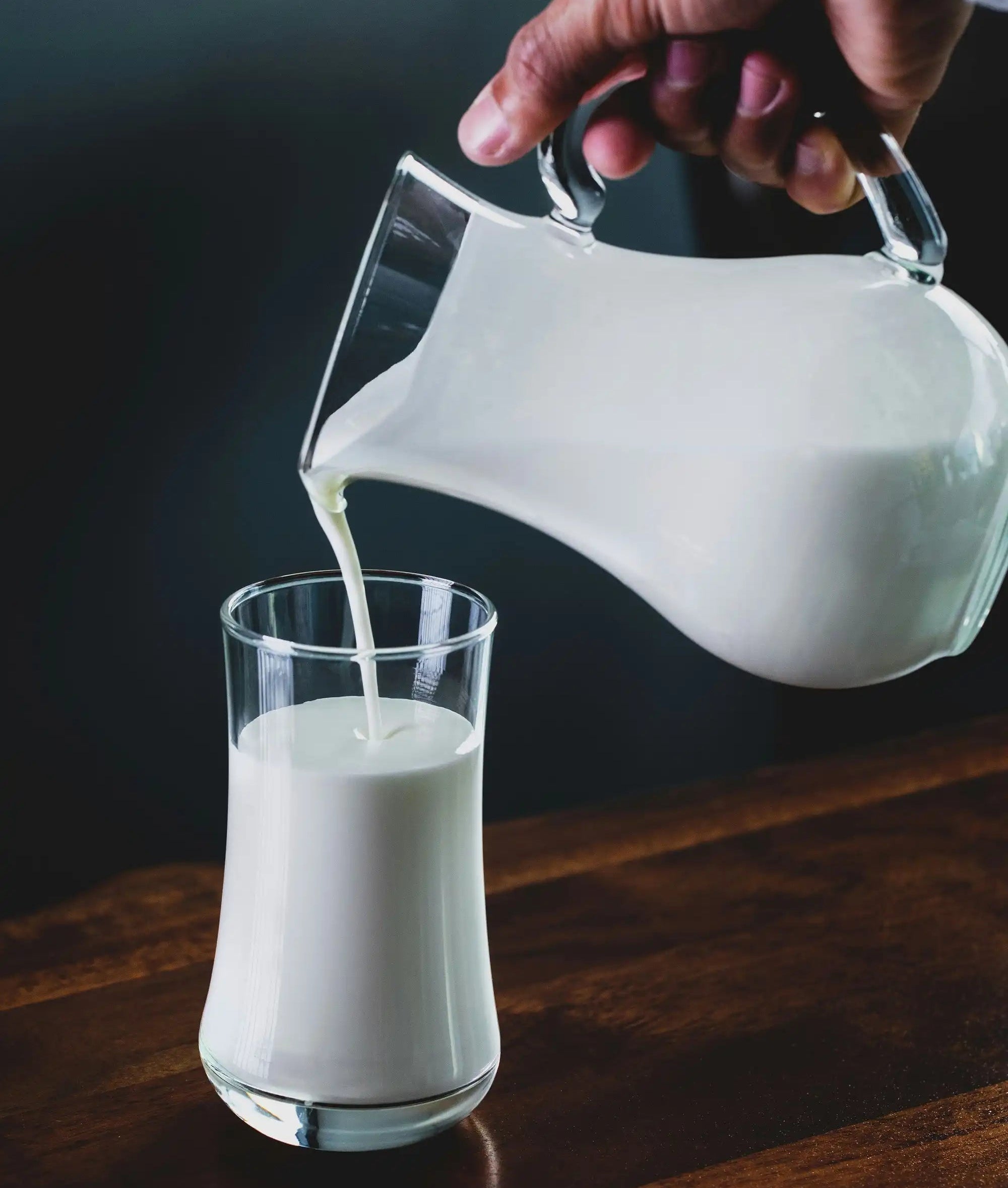 Person pouring milk from a pitcher into a glass on a wooden surface.